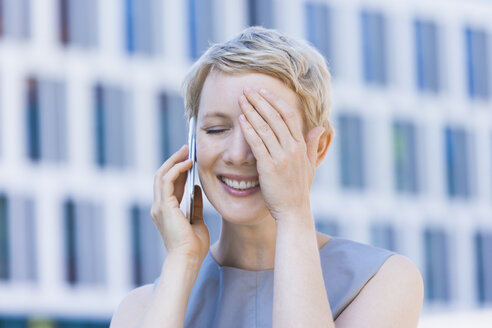 Lächelnde blonde Frau, die mit einem Smartphone telefoniert und ein Auge mit der Hand bedeckt - TCF004296