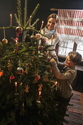Mutter und ihr kleiner Sohn zünden Kerzen an einem Weihnachtsbaum in einem Bauernhaus an - HHF004857