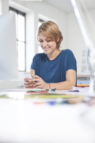 Porträt einer lächelnden jungen Frau, die ein Smartphone an ihrem Schreibtisch in einem kreativen Büro benutzt, lizenzfreies Stockfoto