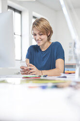 Porträt einer lächelnden jungen Frau, die ein Smartphone an ihrem Schreibtisch in einem kreativen Büro benutzt - RBF001805