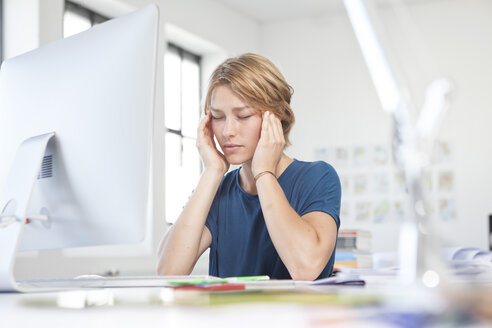 Porträt einer jungen Frau mit Kopfschmerzen an ihrem Schreibtisch in einem kreativen Büro - RBF001808