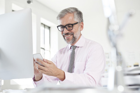 Porträt eines lächelnden Mannes, der ein Smartphone an seinem Schreibtisch in einem Büro benutzt, lizenzfreies Stockfoto