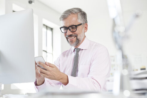 Porträt eines lächelnden Mannes, der ein Smartphone an seinem Schreibtisch in einem Büro benutzt - RBF001824