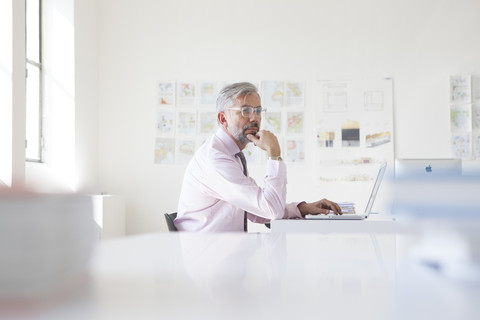 Nachdenklicher Geschäftsmann mit Laptop an seinem Schreibtisch in einem Büro, lizenzfreies Stockfoto