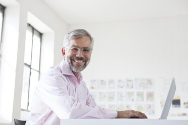 Portrait of smiling businessman with laptop at his desk in an office - RBF001888