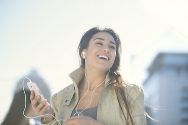Portrait of laughing woman with smartphone and earphones listening music - ZEF000773