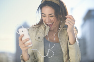 Portrait of laughing woman with smartphone and earphones listening music - ZEF000777