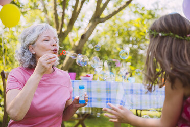 Großmutter bläst Seifenblasen für ein im Garten spielendes Mädchen - MFF001276