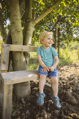 Lächelnder Junge sitzt auf einer Bank unter einem Baum, lizenzfreies Stockfoto