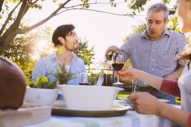 Senior man pouring red wine into glasses of couple having dinner in garden - MFF001328