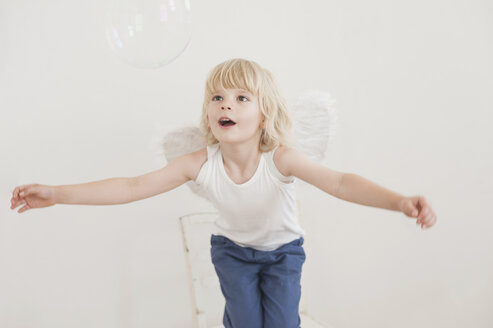 Portrait of gaping little boy with angle wings watching a soap bubble - MJF001341