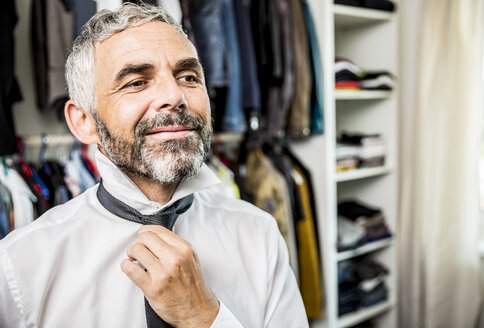 Porträt eines lächelnden Geschäftsmannes, der eine Krawatte in seinem begehbaren Kleiderschrank bindet - MBEF001196
