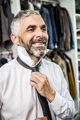 Porträt eines lächelnden Geschäftsmannes, der eine Krawatte in seinem begehbaren Kleiderschrank bindet - MBEF001197