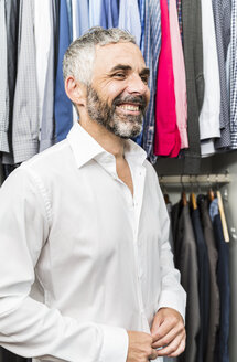 Porträt eines lächelnden Geschäftsmannes, der sein Hemd in seinem begehbaren Kleiderschrank zuknöpft - MBEF001198