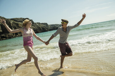 Teenage couple running hand in hand at waterside of the beach - UUF001677