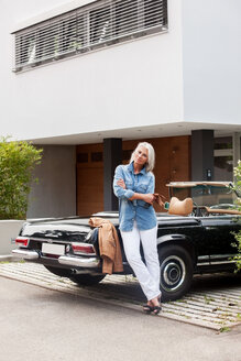 Ältere Frau lehnt sich an ein Cabrio vor ihrem Einfamilienhaus - CHAF000174