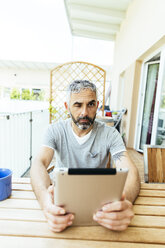Porträt eines Mannes mit digitalem Tablet, der auf seinem Balkon sitzt - MBEF001117