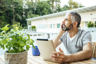 Nachdenklicher Mann mit digitalem Tablet auf seinem Balkon sitzend - MBEF001118