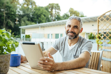 Porträt eines lächelnden Mannes, der auf seinem Balkon sitzt und ein digitales Tablet benutzt - MBEF001119