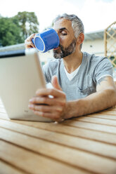 Portrait of man sitting on his balcony drinking coffee using digital tablet - MBEF001120