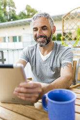 Porträt eines lächelnden Mannes, der auf seinem Balkon sitzt und ein digitales Tablet benutzt - MBEF001121