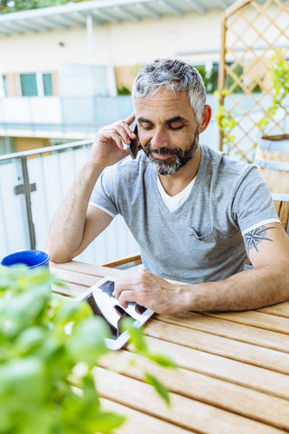 Porträt eines Mannes, der auf seinem Balkon sitzt und mit seinem Smartphone telefoniert, während er ein digitales Tablet benutzt, lizenzfreies Stockfoto