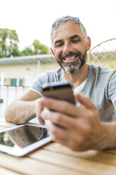 Porträt eines lächelnden Mannes, der auf seinem Balkon sitzt und sein Smartphone mit einem digitalen Tablet benutzt - MBEF001122