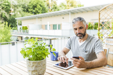 Porträt eines Mannes, der auf seinem Balkon sitzt und ein Smartphone und ein digitales Tablet benutzt - MBEF001229