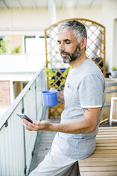 Mann auf seinem Balkon mit Smartphone und einer Tasse Kaffee - MBEF001149