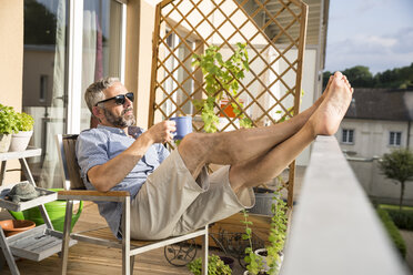 Mann mit Sonnenbrille entspannt sich auf seinem Balkon mit einer Tasse Kaffee - MBEF001129