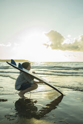 Jugendlicher, der mit seinem Surfbrett am Ufer des Meeres hockt - UUF001722