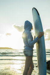 Teenager-Mädchen steht am Strand mit ihrem Surfbrett und schaut auf den Horizont - UUF001724