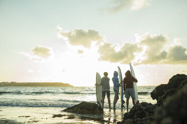 Three teenagers standing on the beach with their surfboards looking at horizon - UUF001725