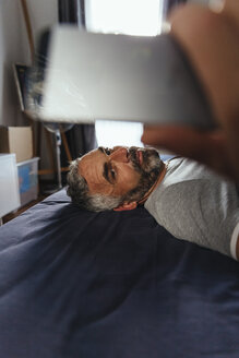 Serious looking man lying on his bed taking a selfie with his smartphone - MBEF001146