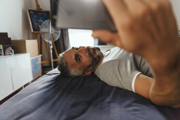 Ernst dreinblickender Mann, der auf seinem Bett liegt und ein Selfie mit seinem Smartphone macht - MBEF001217