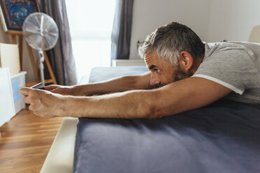 Mann liegt auf seinem Bett und macht ein Selfie mit seinem Smartphone - MBEF001218