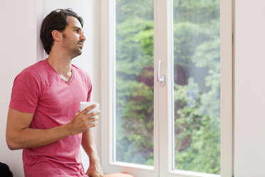 Schläfriger junger Mann mit einer Tasse Kaffee, der aus dem Fenster schaut - JUNF000044