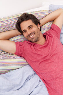 Porträt eines lächelnden jungen Mannes mit den Händen hinter dem Kopf auf dem Bett liegend - JUNF000047