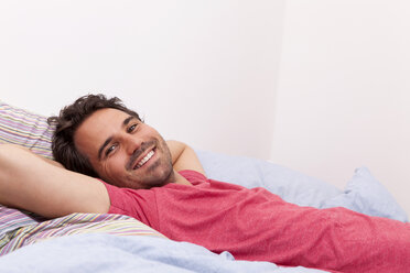 Portrait of smiling young man with hands behind head lying on his bed - JUNF000048