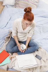 Junge Frau sitzt auf ihrem Bett mit einer Tasse Kaffee und schaut auf ihren Laptop - JUNF000064