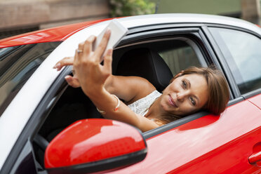 Porträt einer lächelnden jungen Frau, die in ihrem Auto sitzt und ein Selfie mit ihrem Smartphone macht - JUNF000072