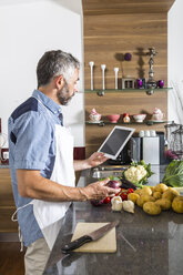 Austria, Man in kitchen with digital tablet preparing food - MBEF001265