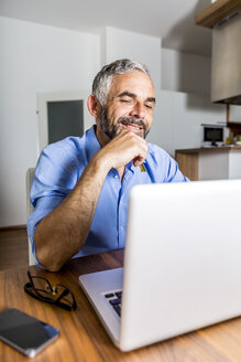 Porträt eines lächelnden Geschäftsmannes, der im Büro zu Hause mit einem Laptop arbeitet - MBEF001292