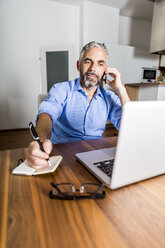 Porträt eines Geschäftsmannes, der mit seinem Smartphone telefoniert und sich Notizen macht - MBEF001294