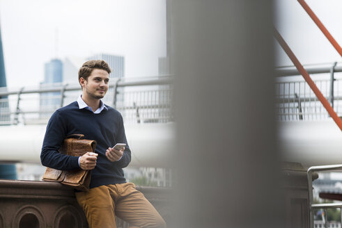 Deutschland, Hessen, Frankfurt, lächelnder junger Mann mit Aktentasche und Smartphone auf einer Brücke stehend - UUF001817