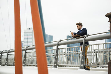 Deutschland, Hessen, Frankfurt, junger Mann steht auf einer Brücke und benutzt sein Smartphone - UUF001818