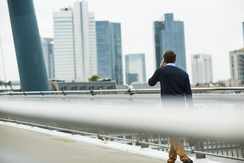 Deutschland, Hessen, Frankfurt, junger Geschäftsmann geht auf einer Brücke und telefoniert mit seinem Smartphone - UUF001819