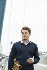 Deutschland, Hessen, Frankfurt, Porträt eines jungen Geschäftsmannes auf einer Brücke mit Aktentasche und Smartphone - UUF001821