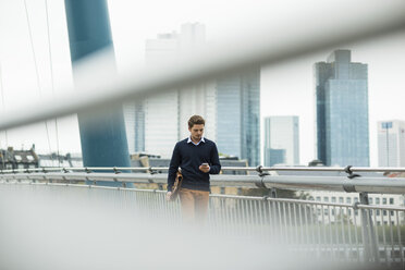 Germany, Hesse, Frankfurt, young businessman walking on a bridge using his smartphone - UUF001822