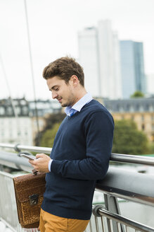 Deutschland, Hessen, Frankfurt, junger Geschäftsmann steht auf einer Brücke und benutzt sein Smartphone - UUF001824